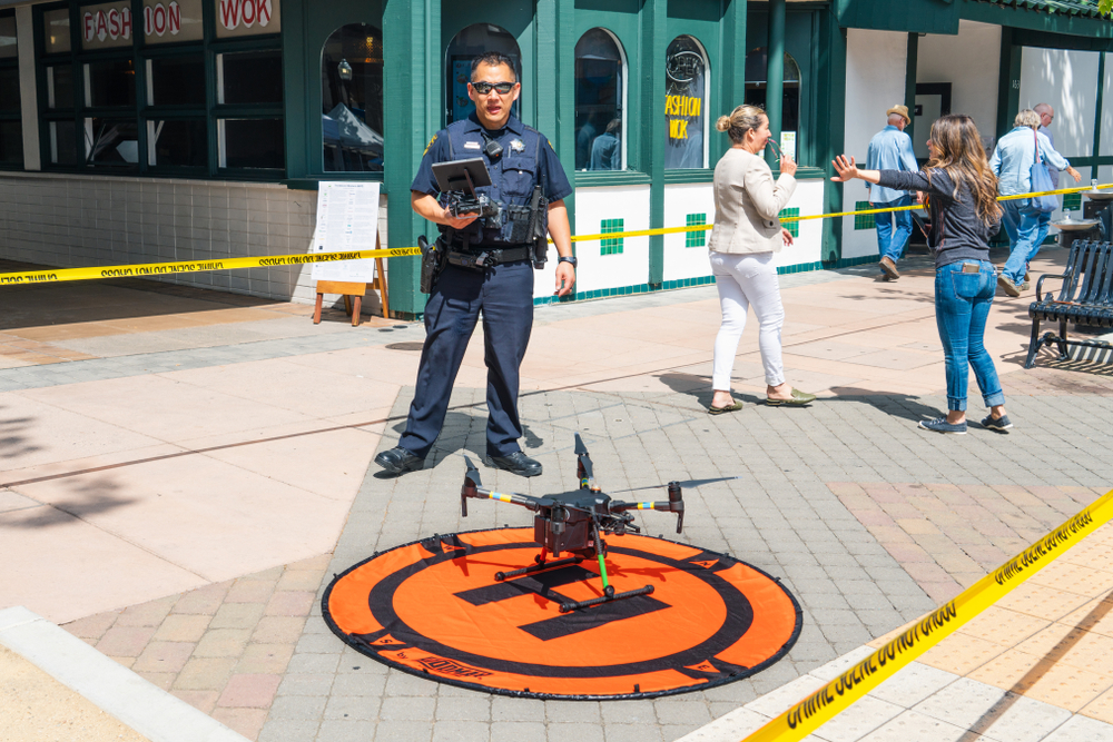 A law enforcement officer standing behind an aerial drone.  The drone is on the ground on an orange landing pad on a city street with by-passers in the background. 