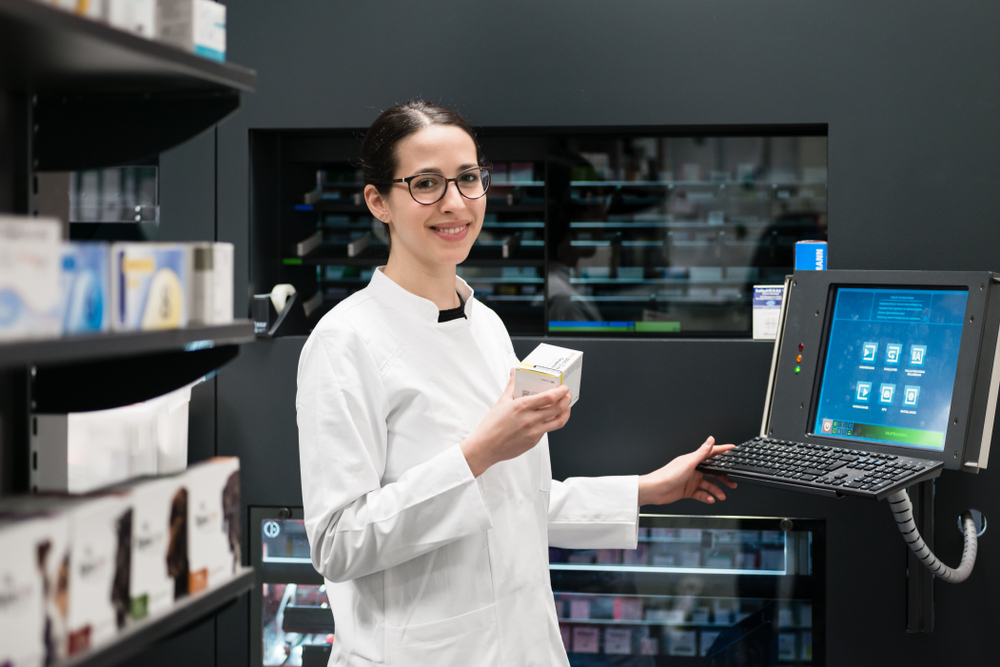 Modern pharmacy. A young woman wearing eyeglasses smiles as she holds a box of medicine. She is wearing a white labcoat. Her other hand touches the keyboard of a computer. Shelves of medications in the background.